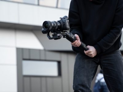 Professional videographer in black hoodie holding professional camera on 3-axis gimbal stabilizer. Filmmaker making a great video with a professional cinema camera. Cinematographer. Professional videographer in black hoodie holding professional camera on 3-axis gimbal stabilizer. Filmmaker making a great video with a professional cinema camera. Cinematographer